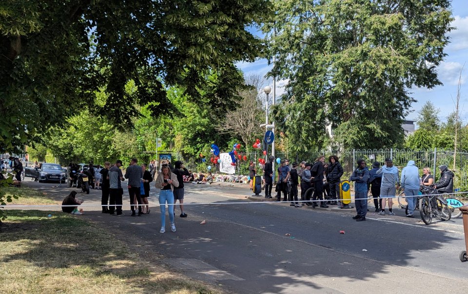 People gathered at a roadside memorial with balloons and flowers.