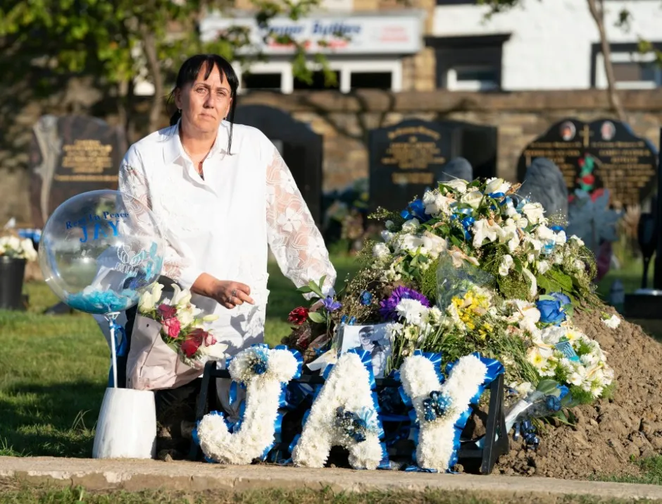 Woman mourning at a gravesite with floral tributes spelling "JAY".
