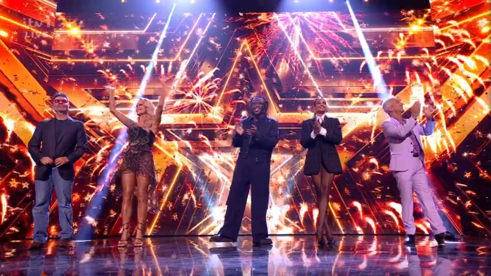 Screenshot of four judges on a stage with a firework background.