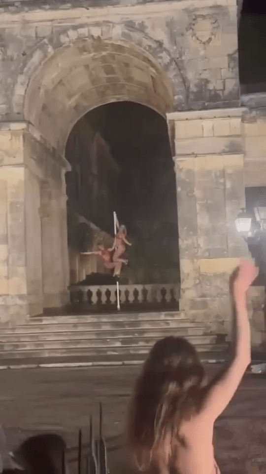 Two pole dancers performing on a pole in front of a stone archway.