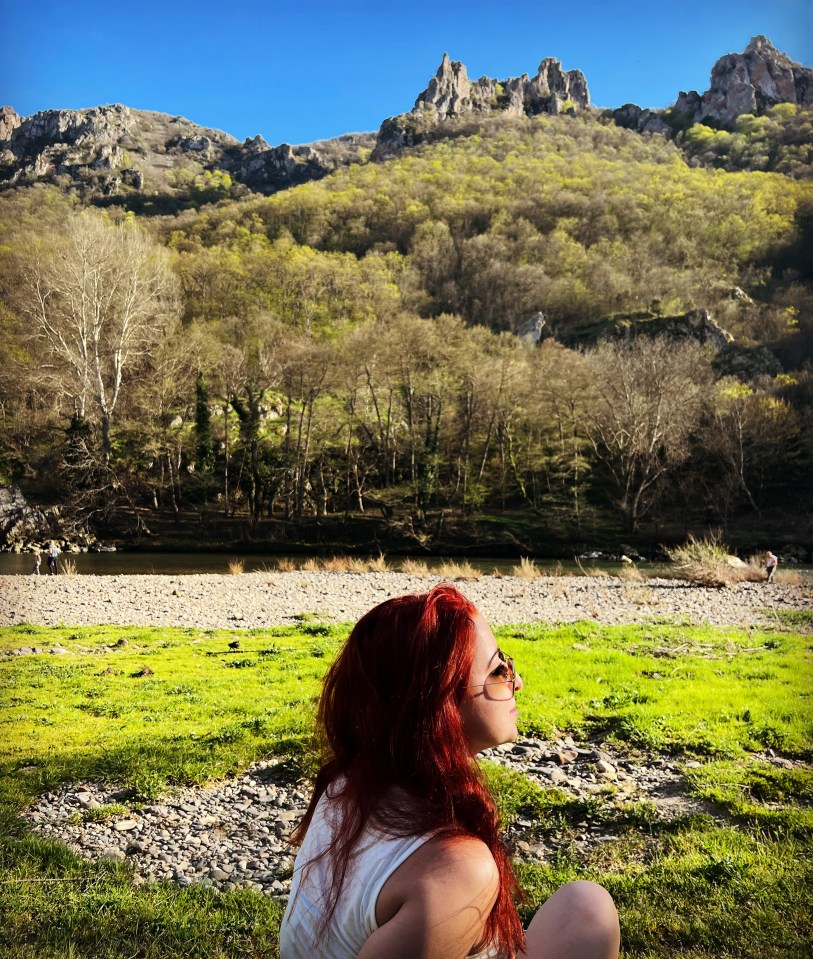 Woman with red hair sitting outdoors, looking at a rocky mountain landscape.