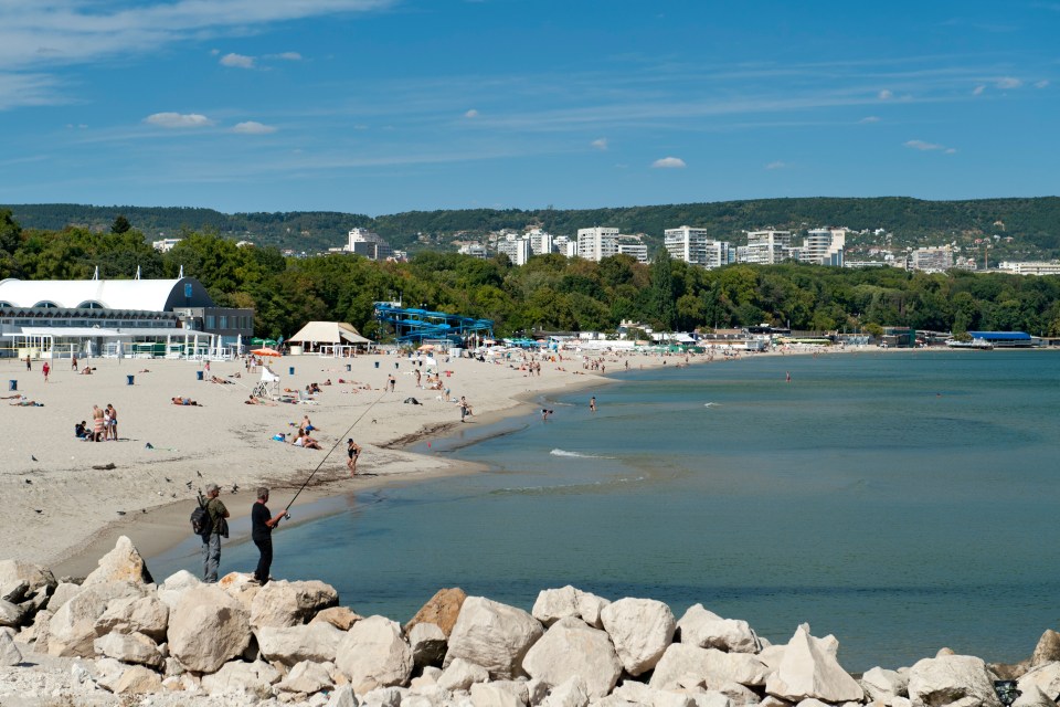 Beach scene in Varna, Bulgaria, with people swimming and sunbathing, and a man fishing from rocks.