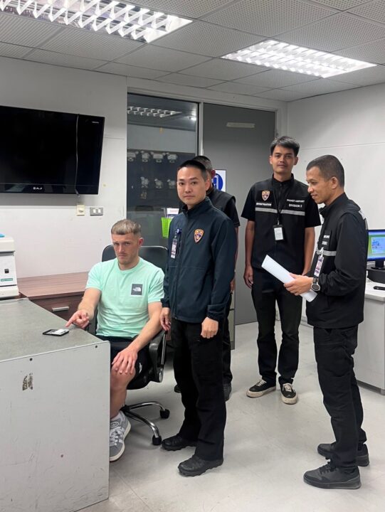 A man sits at a desk being questioned by Thai immigration officers.
