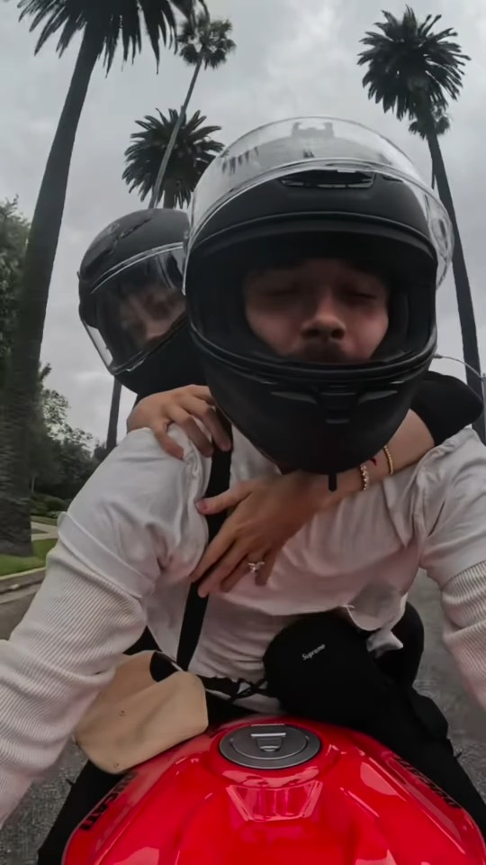 A person wearing a motorcycle helmet hugs another person also wearing a helmet while sitting on a red motorcycle.