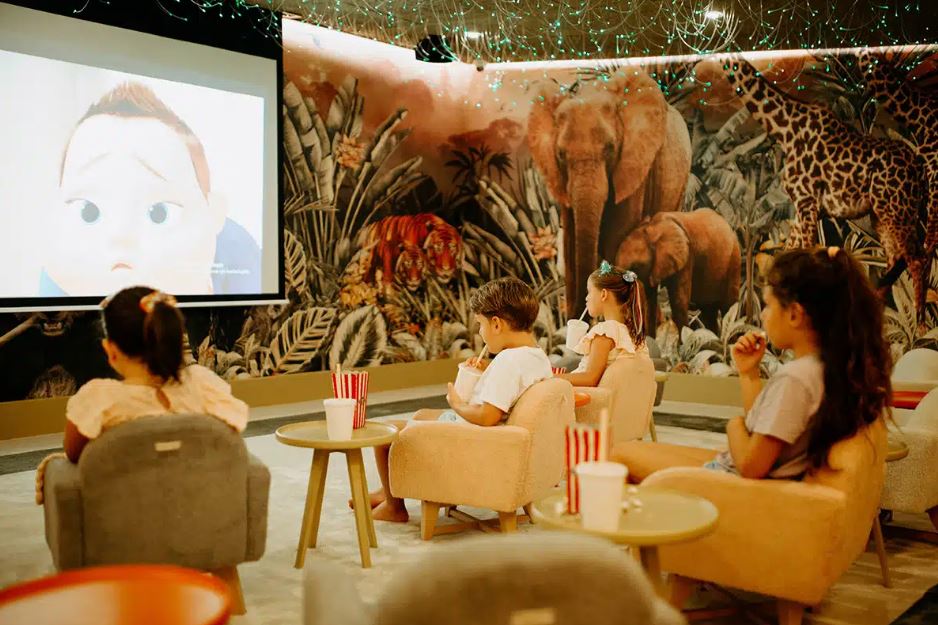 Children watching a movie in a theater with a jungle mural.