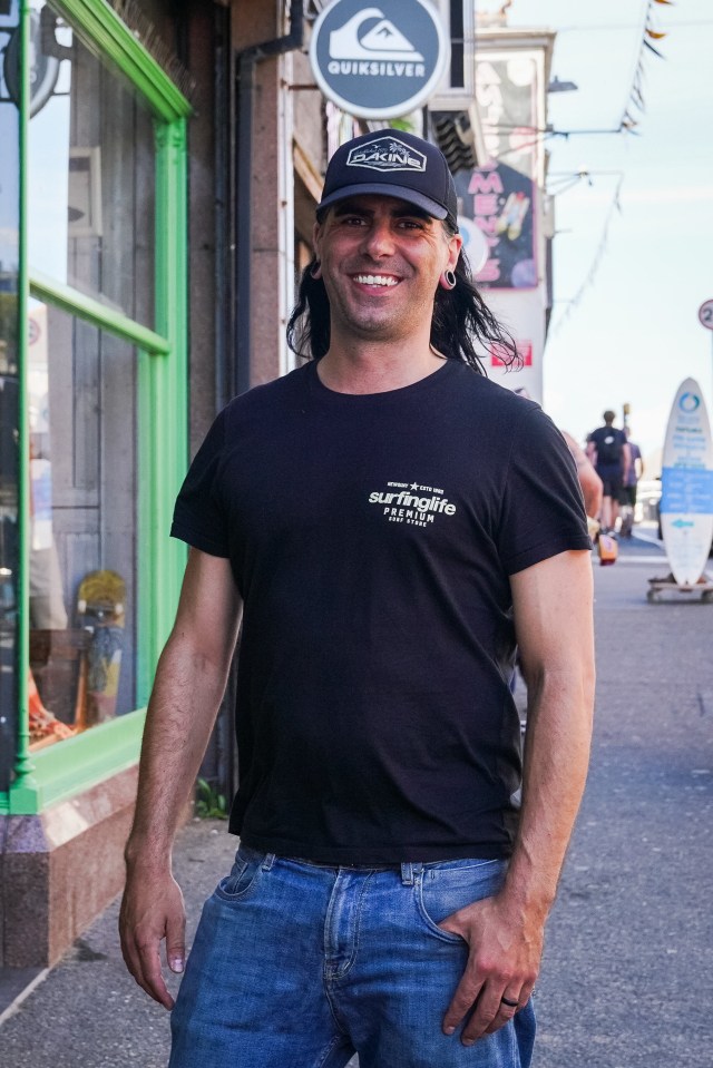 A man with long hair wearing a black t-shirt and jeans smiles for the camera.