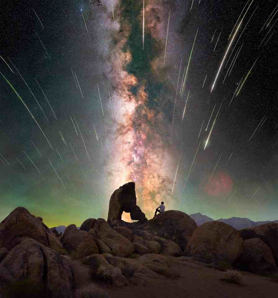 Person sitting on rocks under a night sky filled with the Milky Way and numerous meteors.