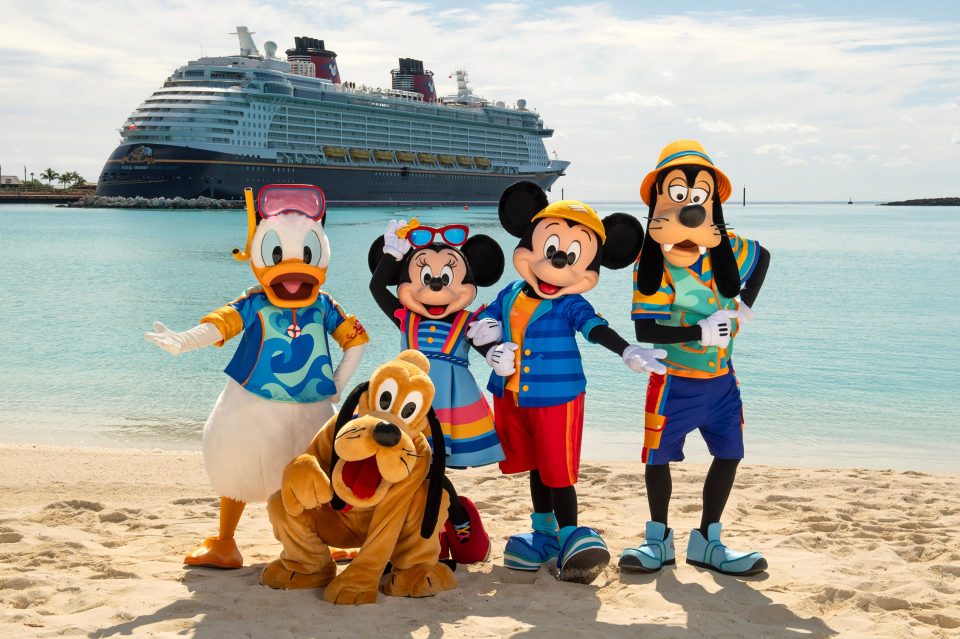 Mickey Mouse, Minnie Mouse, Goofy, and Donald Duck in beach attire on a beach with a Disney cruise ship in the background.