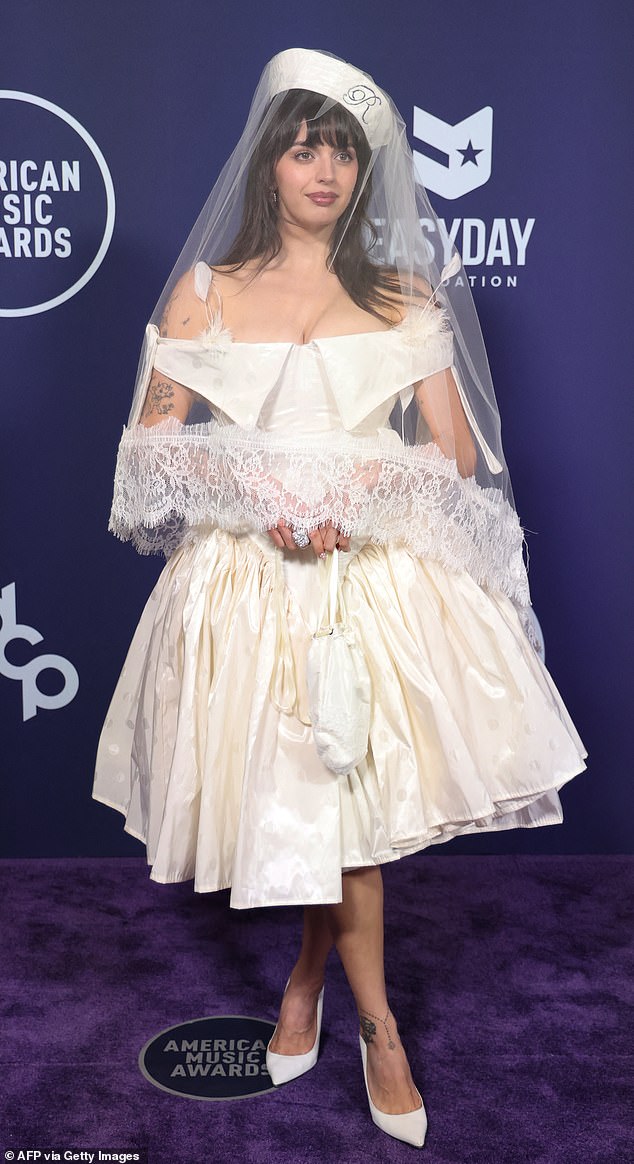 Singer Rebecca Black hit the red carpet in a strange white gown that resembled a wedding dress. It contained dramatic, off-the-shoulder sleeves, feathers, and a large puffy skirt