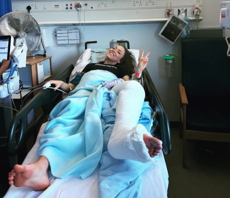 Woman in hospital bed with leg cast, giving peace sign.