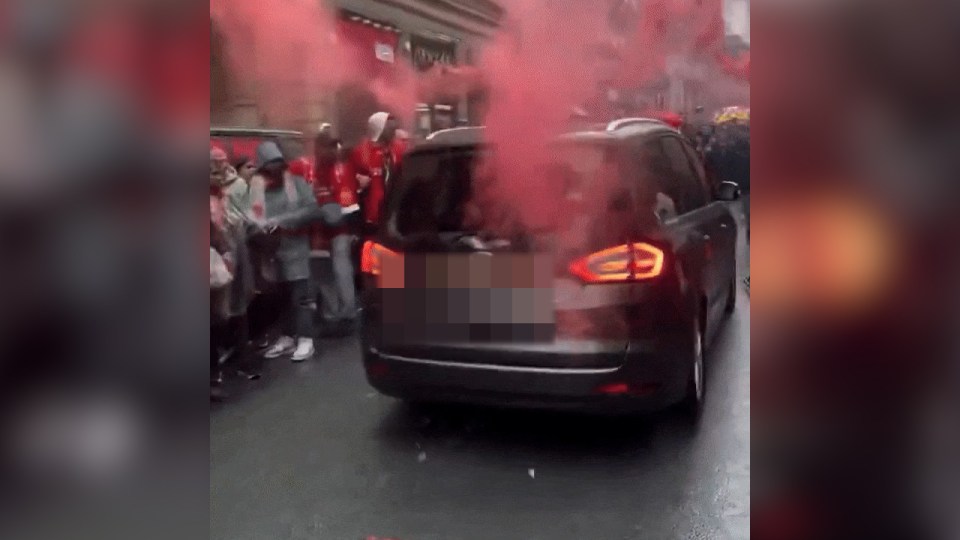 A dark-colored minivan driving through a crowd of Liverpool fans amidst red smoke.