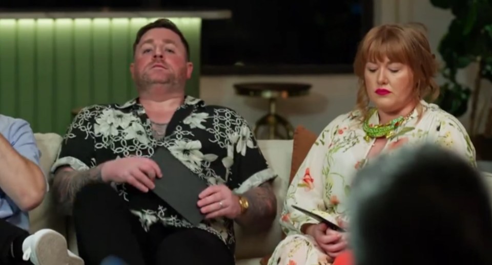 A man and a woman sit on a couch, looking serious.