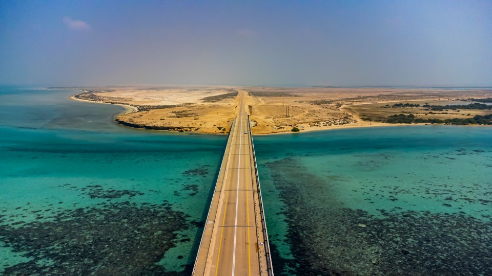 Aerial view of a bridge connecting a desert island to the mainland, with turquoise water visible below.