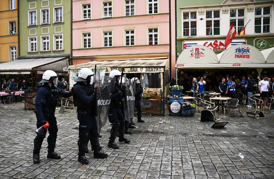 Police officers in riot gear stand outside a restaurant where overturned chairs are visible.