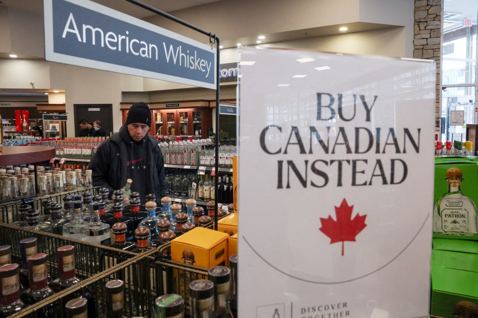 A customer holds a bottle of liquor in a store with a sign that says "Buy Canadian Instead".