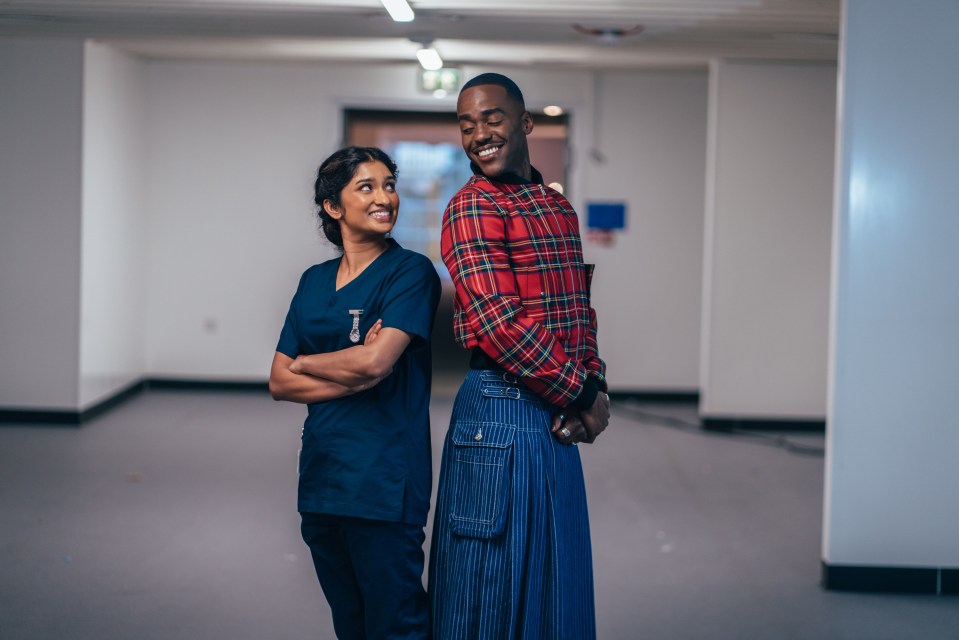A doctor and another person stand in a hallway, smiling at each other.
