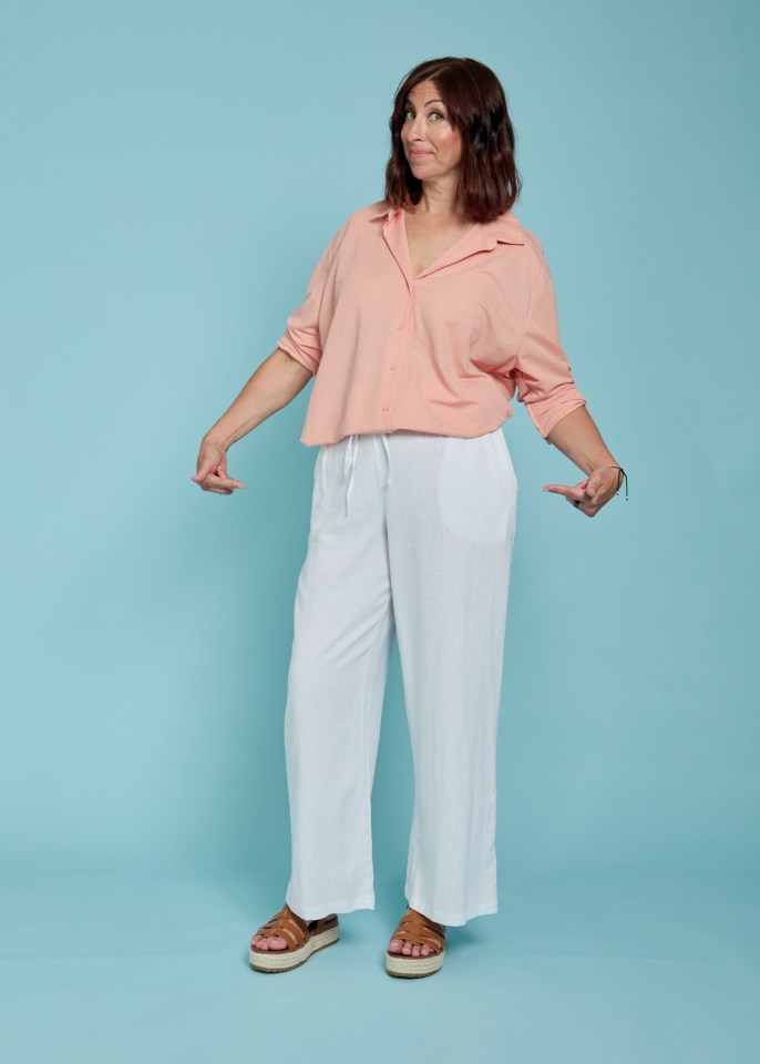 Woman in peach shirt and white pants gesturing towards her outfit.