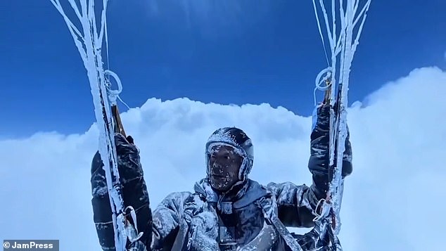 The paraglider can be seen dangling above the clouds - visibly frozen with his hair, beard, and protective gear covered in frost