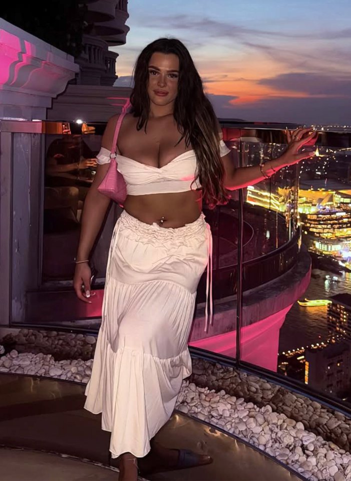 A young woman in a white off-the-shoulder crop top and maxi skirt stands on a rooftop overlooking a city at sunset.