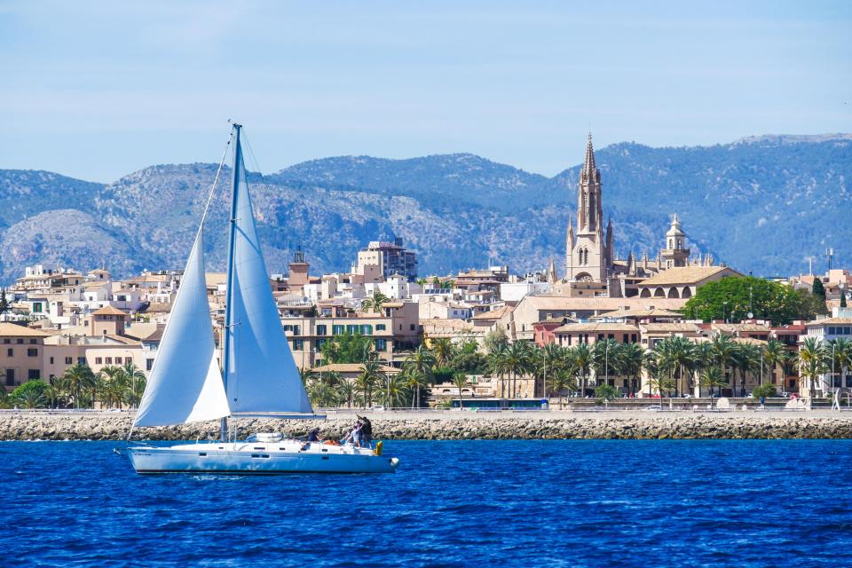 Sailboat in Palma de Mallorca harbor with city and mountains in the background.