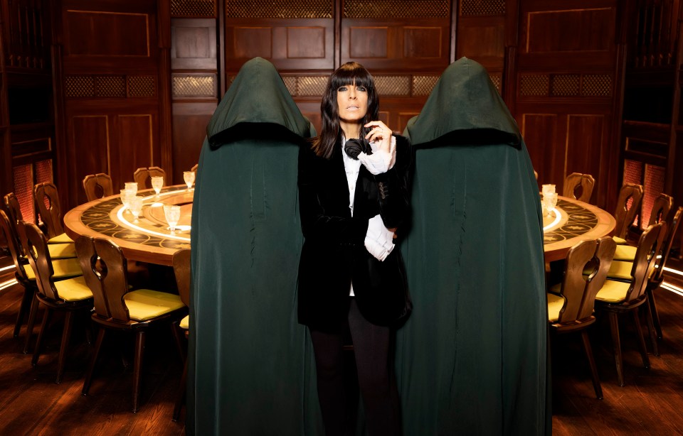 Claudia Winkleman standing between two figures in hooded cloaks at a round table.