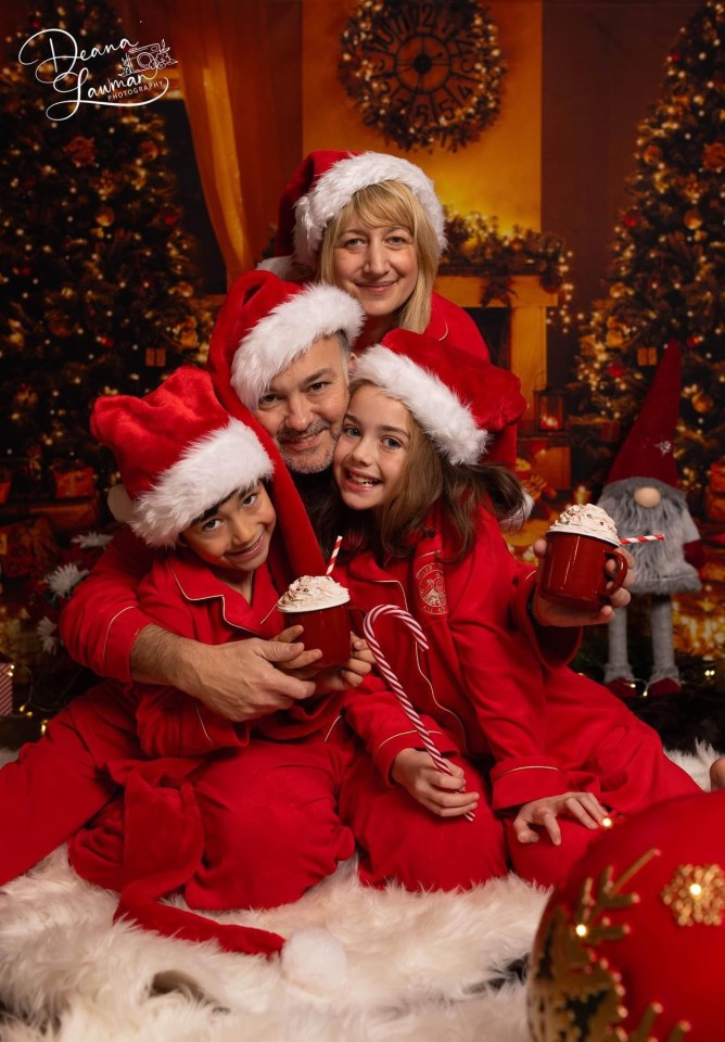 A family of four sitting on a couch, with the father in a wheelchair with an oxygen tank, and the mother on the other side of him holding a young boy on her lap. They are all wearing Santa hats and smiling.