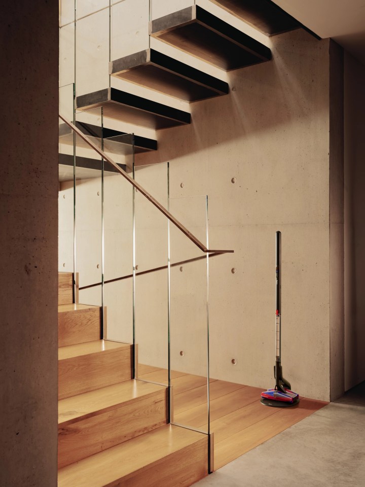 A cordless vacuum cleaner stands on a wooden floor next to a modern staircase with glass railings.