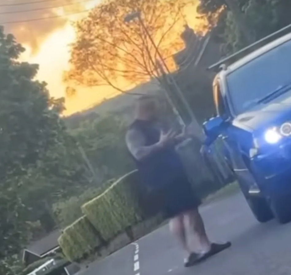 Screenshot of a man standing in the road next to a blue car at sunset.