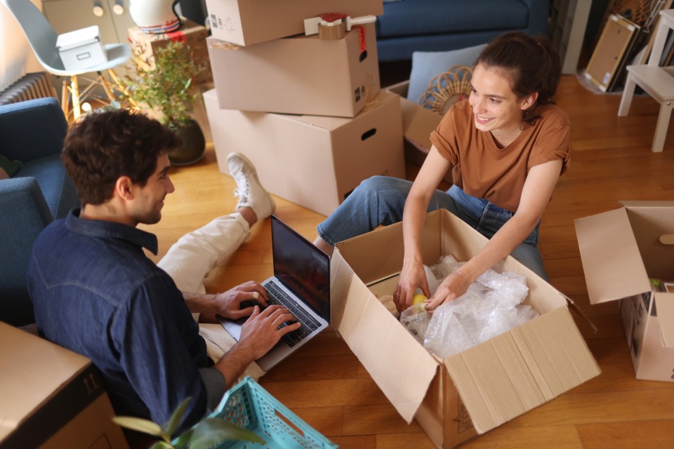 A young couple unpacking boxes in their new apartment, one using a laptop.