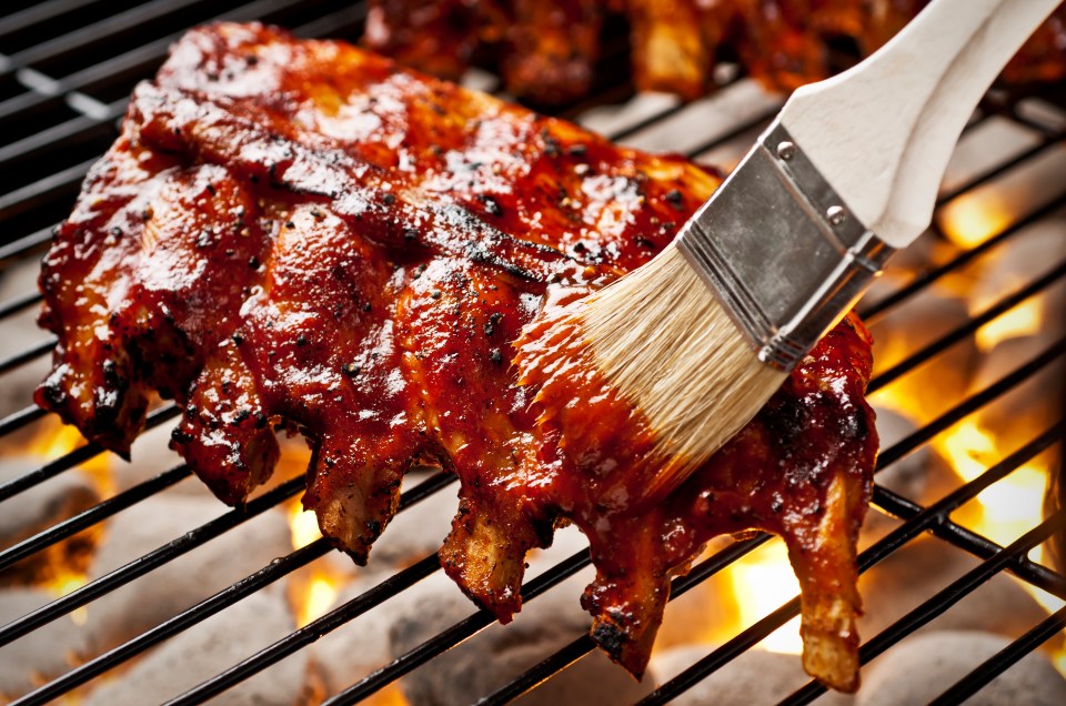 Baby back ribs being basted with barbecue sauce on a grill.