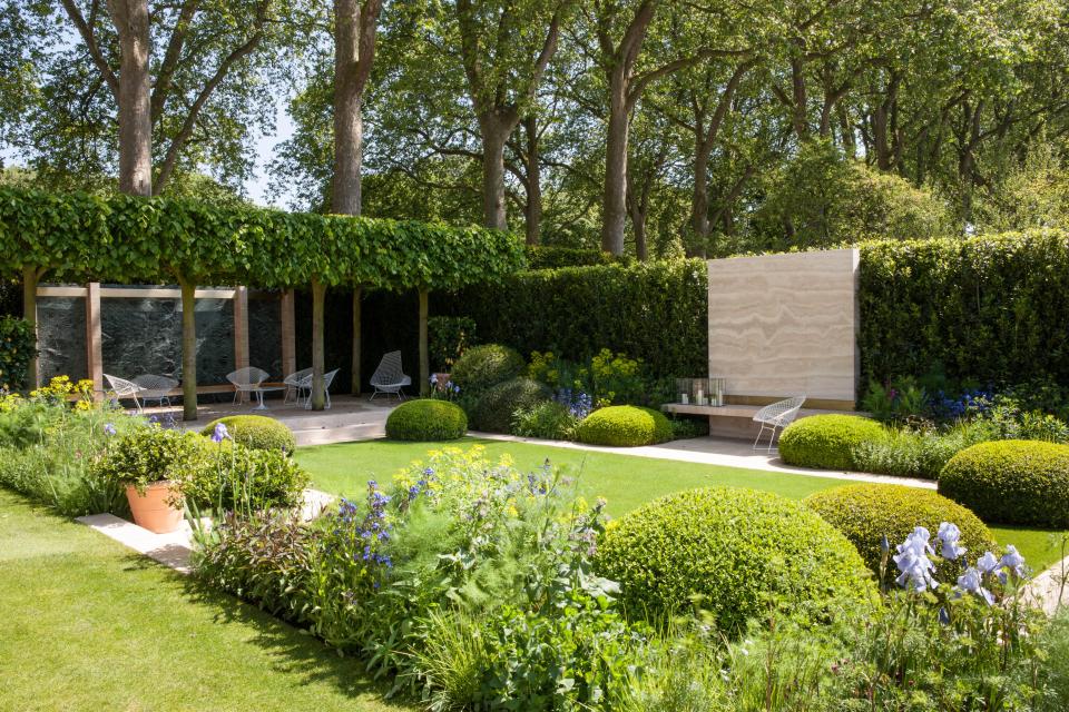 A garden with a stone wall, hedges, and modern chairs.