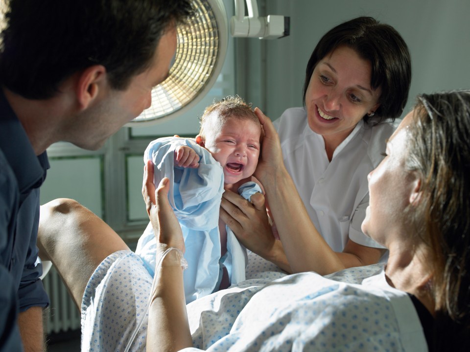 A newborn baby cries as a nurse hands the baby to its parents.