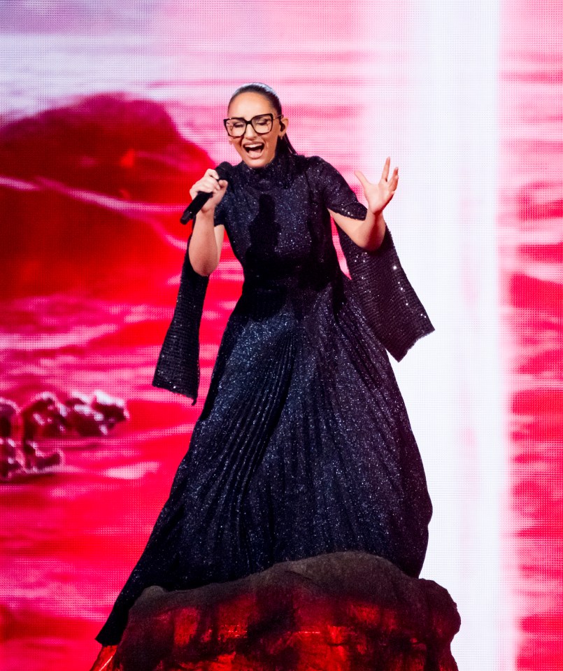A woman in a sparkly black dress sings into a microphone on a stage with a red background.