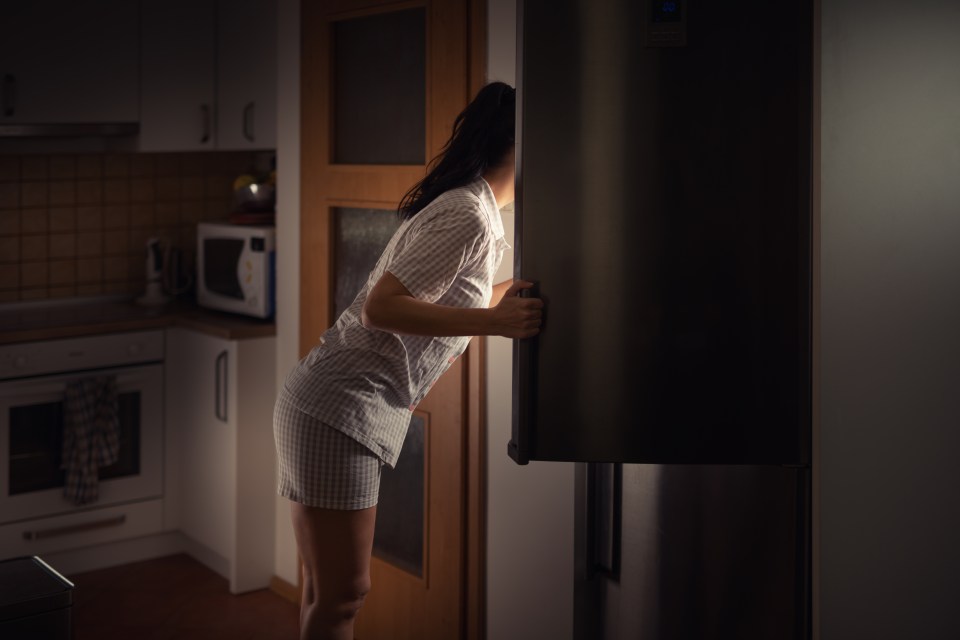 Woman in pajamas looking into a refrigerator at night.
