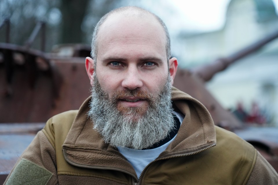 Portrait of a man with a long grey beard, wearing a tan jacket, near destroyed military vehicles.