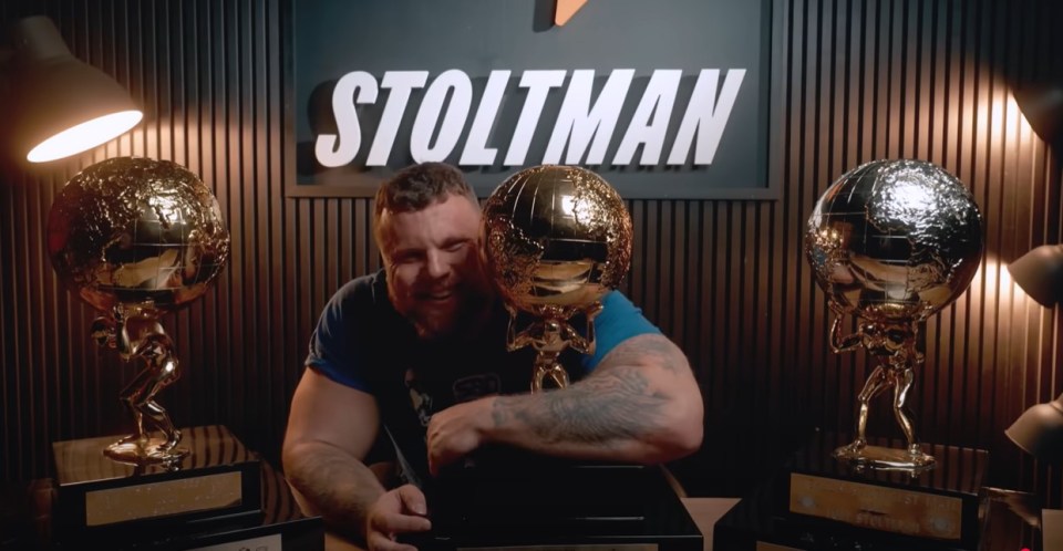 A strongman smiles while holding a golden globe trophy.