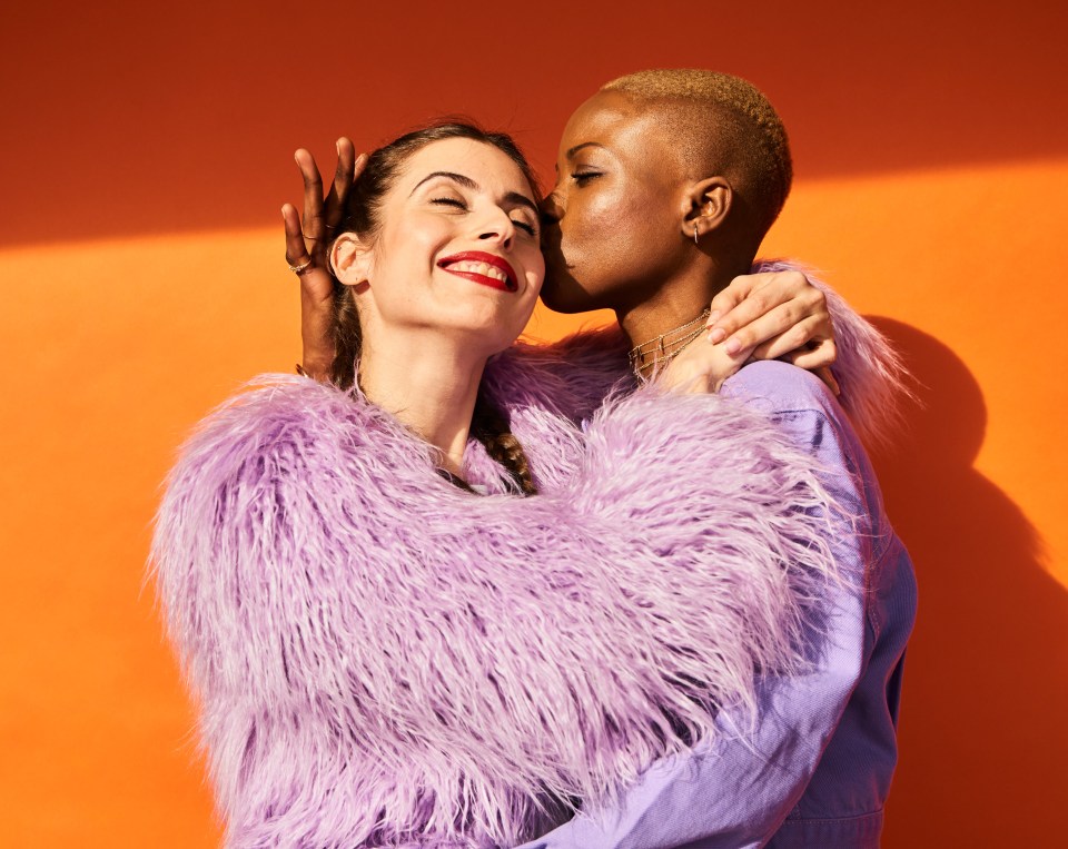 Two women embracing and kissing in front of an orange background.