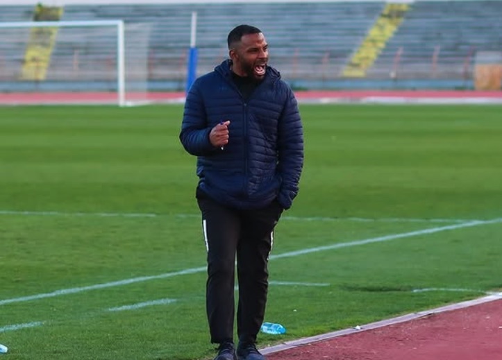 Man in dark blue jacket coaching on soccer field.