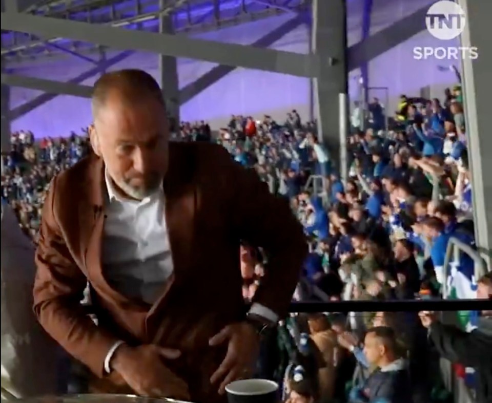 Man in brown jacket sits before a cheering crowd.