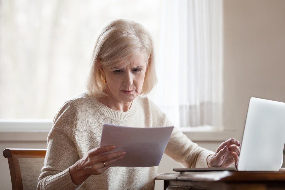A senior woman looking worried while reviewing paperwork and using a laptop.