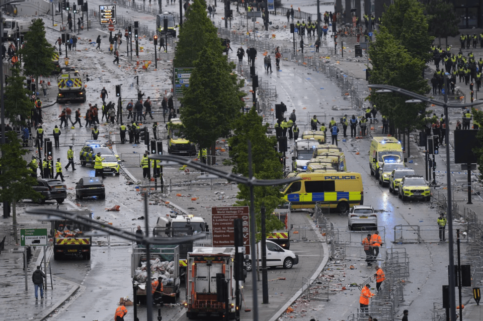A street littered with trash after a large event, with police and cleanup crews present.