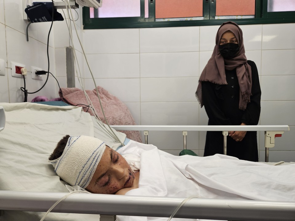 A wounded Palestinian child lies in a hospital bed, with a woman standing nearby.