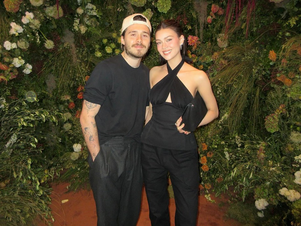 Brooklyn Beckham and Nicola Peltz Beckham posing together in front of a floral wall.