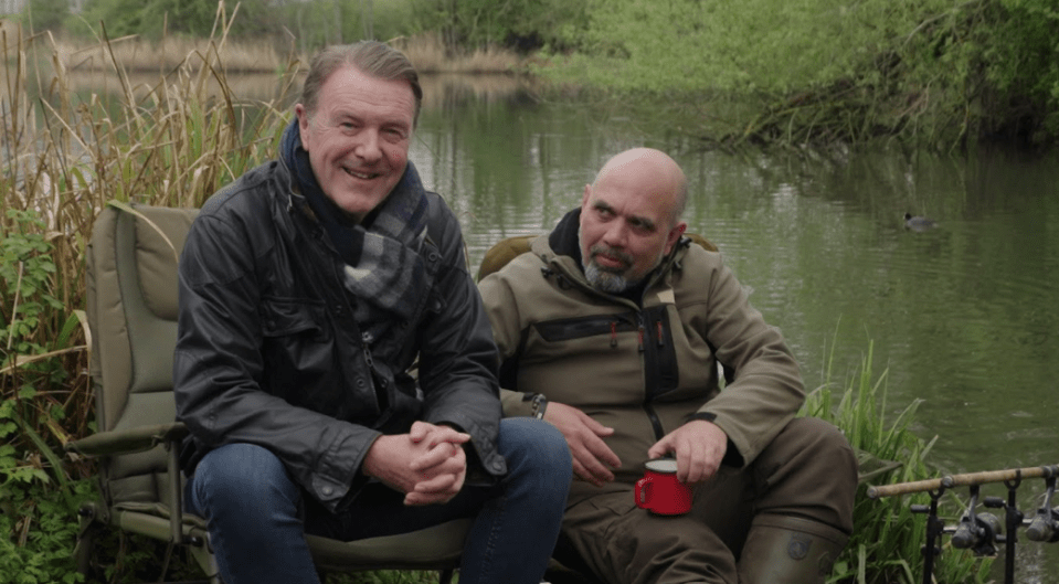 Two men sitting in chairs by a lake, holding mugs.