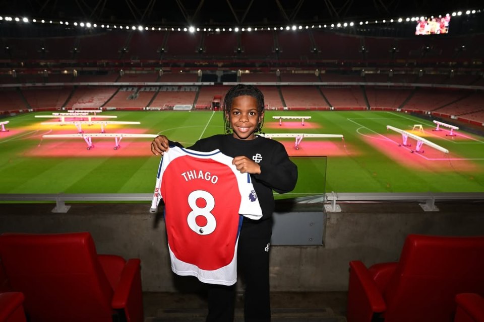 Boy holding Arsenal jersey with "Thiago 8" on the back in an empty stadium.