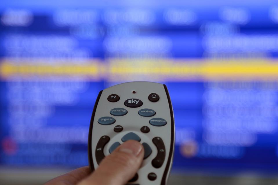 Hand holding a Sky remote control in front of a blurred television screen.