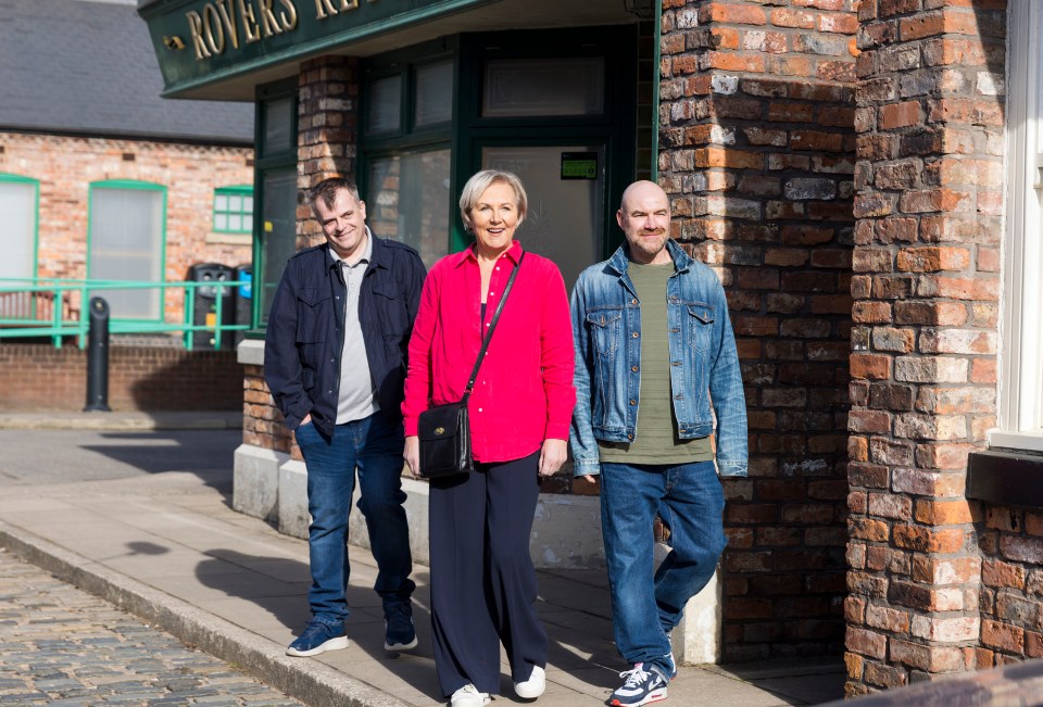 Three Coronation Street actors, Sue Cleaver, Simon Gregson, and Joe Duttine, walking outside a brick building.