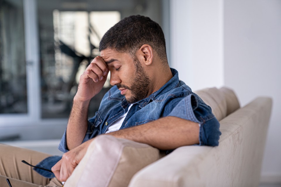A worried man sits on a couch, holding his head.