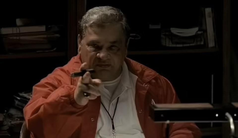 Man in red jacket points while holding a cigar.