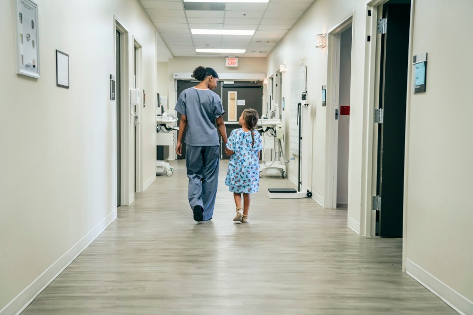 Nurse and young girl holding hands walking down a hospital corridor.